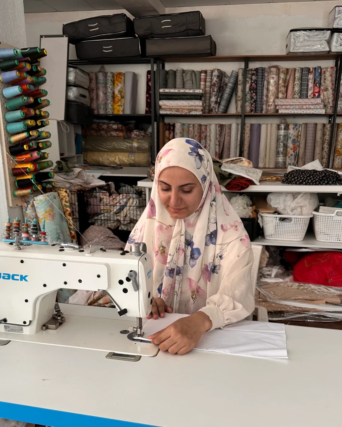 ژنێک بە لەچکێکی گوڵدارەوە قوماشێکی سپی بە مەکینەی دروومان دەدوورێت، لە پشتییەوە بۆبینی دەزوو و ڕەفەی تۆپە قوماش دیارن